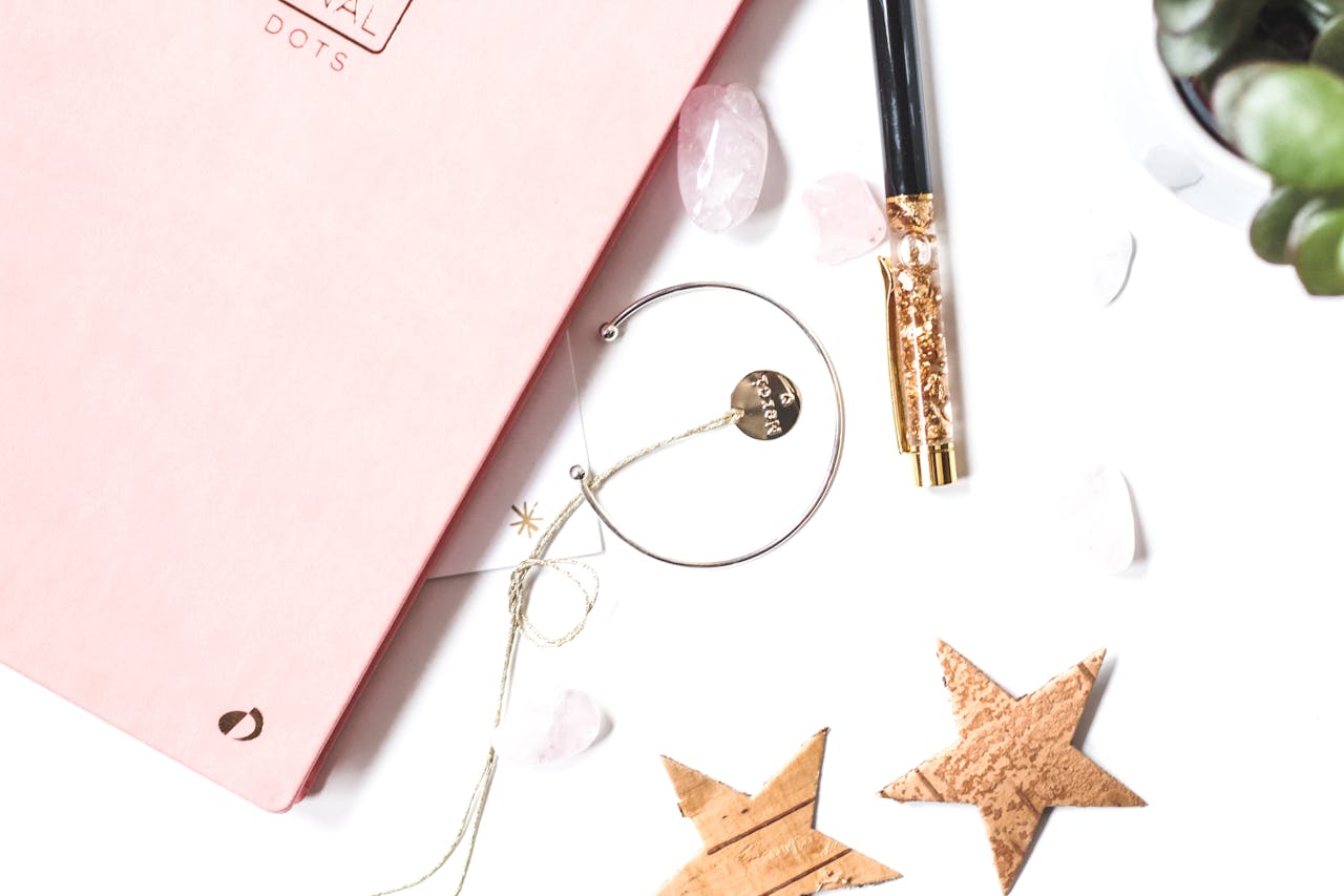 Stylish flat lay featuring a pink journal, gold pen, bracelet, and decorative stars.